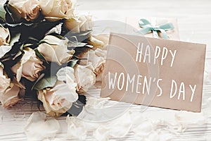 Happy Women`s Day text sign on craft greeting card and white roses bouquet, gift box on wooden background. International womens