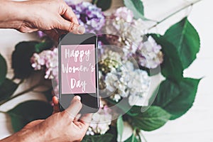 Happy Women`s Day text sign on phone screen in woman hands on background of hydrangea flowers on rustic white wood. Floral