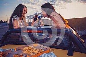 Happy women are smiling, cheering with soda in glass bottles, posing in yellow car with french fries and pizza on its
