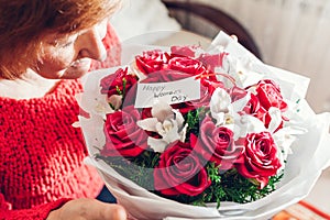 Happy Womens Day, 8 March gift. Senior woman smells bouquet of red roses and orchids flowers at home with card