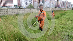 A happy worker in bright overalls mows long grass with a motorcycle mower on the roadside of a residential neighborhood,