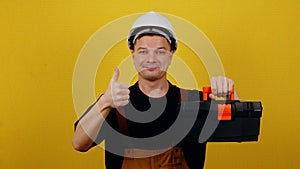 Happy worker in uniform and hard hat holding tool box and showing thumbs up