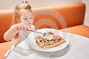 Happy 3 year old girl eat. Breakfast in cafe. Classic Viennese waffles with ice cream, berries and Maple syrup. Table in