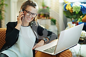 Woman with laptop talking on phone in modern house in sunny day