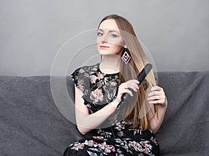 Happy young adorable woman in long dress on grey sofa holding hair styler up at home against grey wall