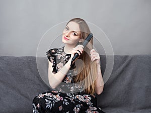 Happy young adorable woman in long dress on grey sofa holding hair styler up at home against grey wall