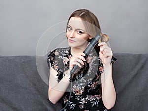 Happy young adorable woman in long dress on grey sofa holding hair styler up at home against grey wall