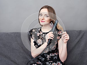 Happy young adorable woman in long dress on grey sofa holding hair styler up at home against grey wall