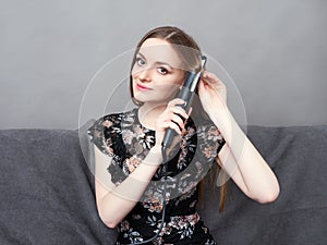 Happy young adorable woman in long dress on grey sofa holding hair styler up at home against grey wall