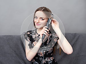Happy young adorable woman in long dress on grey sofa holding hair styler up at home against grey wall