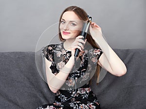 Happy young adorable woman in long dress on grey sofa holding hair styler up at home against grey wall