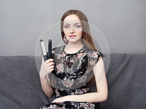 Happy young adorable woman in long dress on grey sofa holding hair styler up at home against grey wall