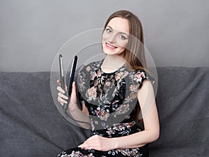 Happy young adorable woman in long dress on grey sofa holding hair styler up at home against grey wall