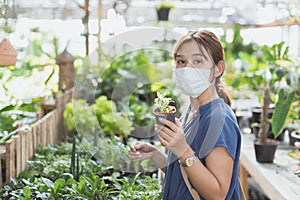 Happy young adult Asian woman with medical face mask holding pot, little tree pot, pot of flowers or plants at greenhouse or