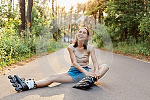 Happy young adult woman in rollerblades, sitting on road, wearing beige t shirt and jeans short, looking directly at camera and
