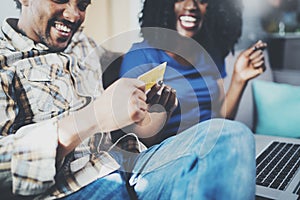 Happy young african american couple sitting on sofa at home and shopping online through mobile computer by credit card