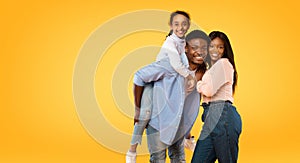 Happy young african american family posing, man giving piggy back ride for his daughter, yellow background, free space