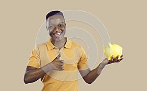 Happy young African American man holding piggy bank, smiling and giving thumbs up