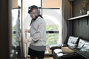 Happy young African American man with wireless headphone having fun cleaning house