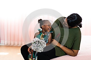 Happy young African American mother having fun with daughter child girl playing and hugging in bedroom at home