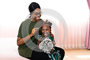 Happy young African American mother having fun with daughter child girl playing and hugging in bedroom at home