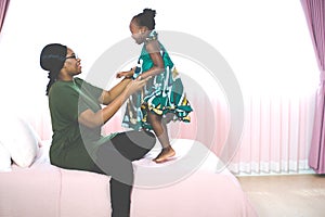 Happy young African American mother having fun with daughter child girl playing and hugging in bedroom at home