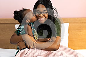 Happy young African American mother having fun with daughter child girl playing and hugging in bedroom at home