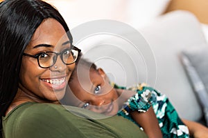 Happy young African American mother having fun with daughter child girl playing and hugging in bedroom at home