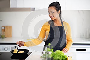 Happy young african american woman in apron frying fish at table with organic vegetables