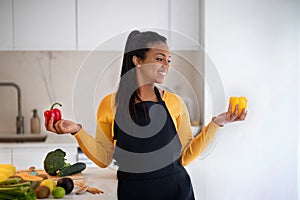 Happy young african american woman in apron hold red and yellow pepper in hands at table with organic vegetables