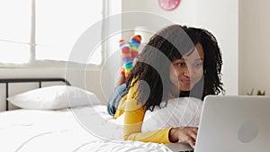Happy young african american woman lying on bed using laptop computer