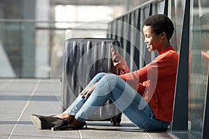 Happy young african american woman sitting on floor in airport terminal with luggage and looking at mobile phone