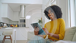 Happy young African American woman sitting on sofa at home using digital tablet.