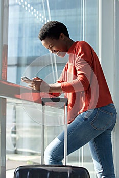Happy young african american woman standing in airport terminal looking at cellphone by window