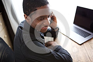 Happy young african businessman sitting at desk with computer and talking on mobile phone