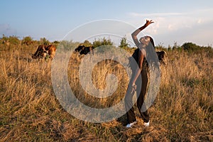 Happy young african girl in black clothes dance among the dry grass field, cows graze on the background