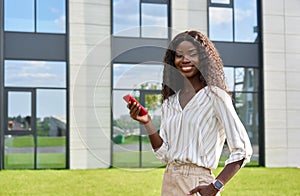 Happy young African lady using mobile phone standing outdoors.
