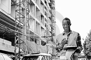 Happy young African man construction worker using phone while holding clipboard at building site