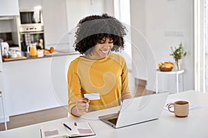 Happy young African woman customer holding credit card using laptop in kitchen.