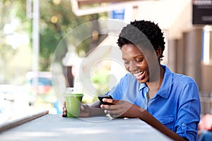 Happy young african woman sitting at outdoor cafe using mobile phone