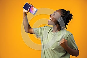 Happy young Afro American woman listening to music in headphones against yellow background