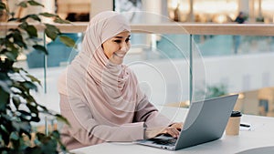 Happy young arab businesswoman sitting at office desk typing on laptop working on internet checking new app smiling