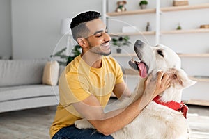 Happy young Arab man stroking his adorable dog in living room. Human animal friendship concept