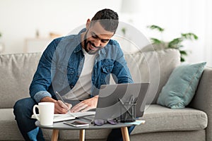 Happy young Arab man taking notes while working on laptop computer at home office, copy space