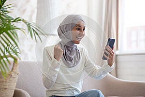 Happy young Arab woman in hijab sitting on sofa at home and using mobile phone. looks at the screen and makes a victory