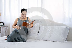 Happy young Asia pregnant woman holding and reading red book and sitting on white bed