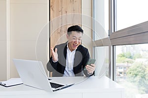 Happy young Asian business man in office looking at mobile phone, with emotion winner or win, financial stock sports betting