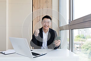 Happy young Asian business man in office looking at mobile phone, with emotion winner or win, financial stock sports betting