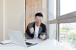 Happy young Asian business man in office looking at mobile phone, with emotion winner or win, financial stock sports betting.