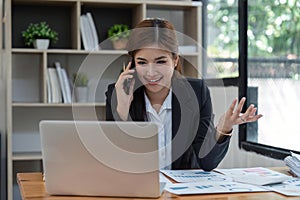 Happy young asian business woman talking on the mobile phone and smiling while sitting at her working place in office